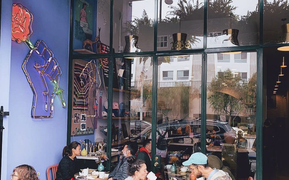 Bloom & Plume Coffee storefront — a neon rose-and-fist sign on the periwinkle wall beside the front window, with guests at orange café chairs